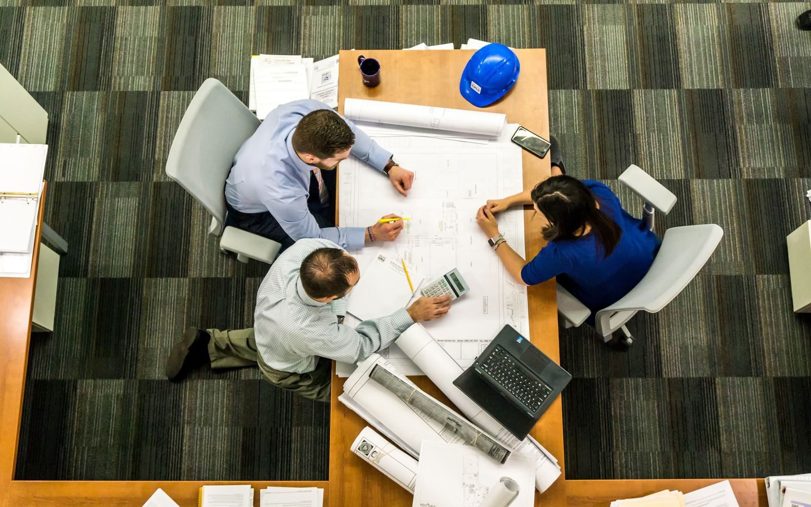 Three people reviewing rolled building plans around a table with a hard hat, calculator and laptop, the plan assessment work that stays with the building surveyor