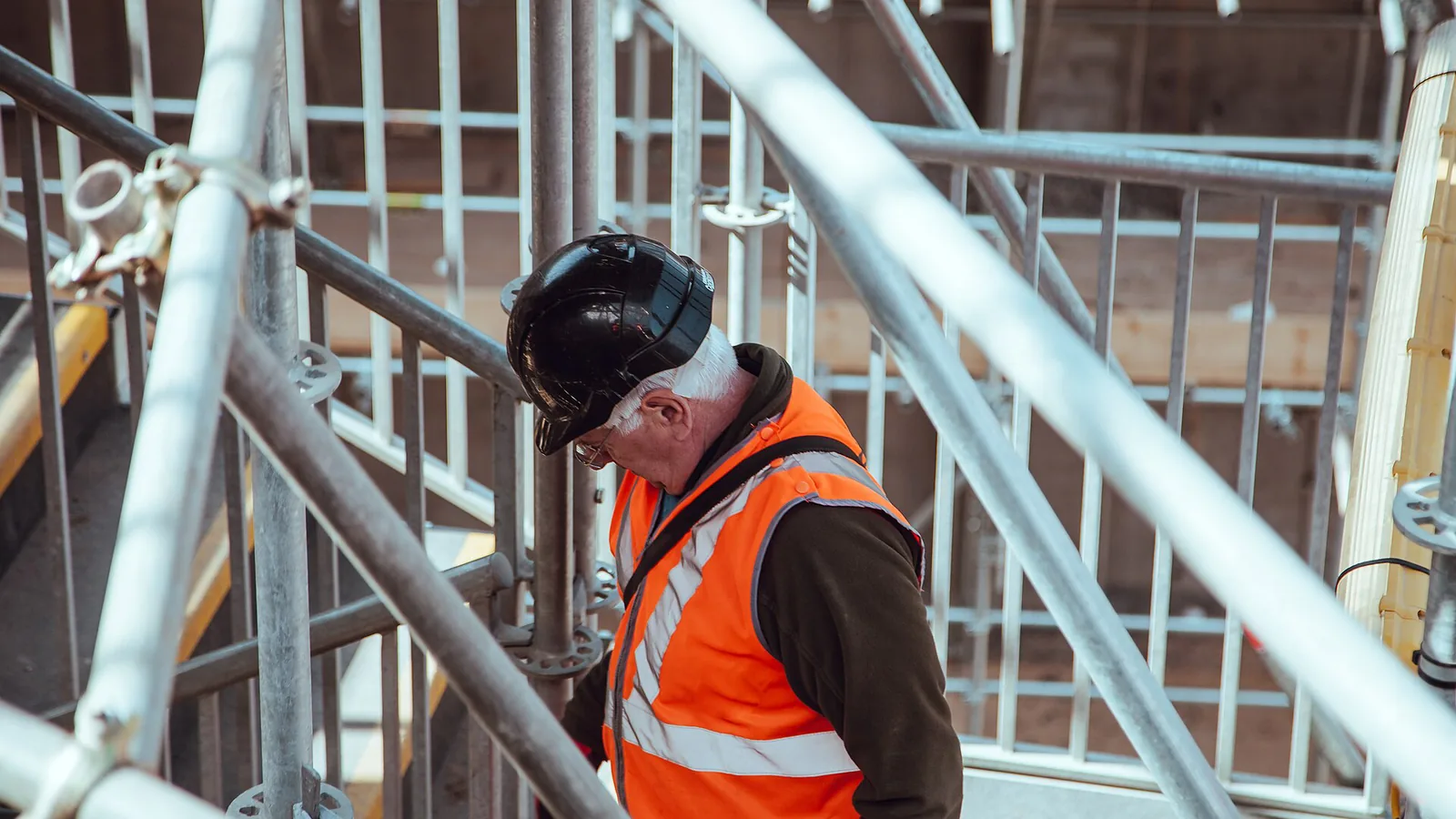 A building surveyor in a hard hat and high-visibility vest on a scaffolded stair, doing the judgment work building surveyor automation cannot replace