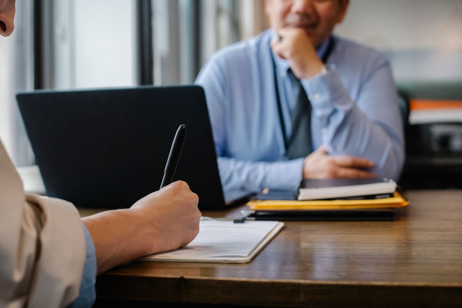 Accountant taking notes at a desk while a client in a blue shirt listens across a laptop and folders, the advice work accounting compliance automation leaves with people
