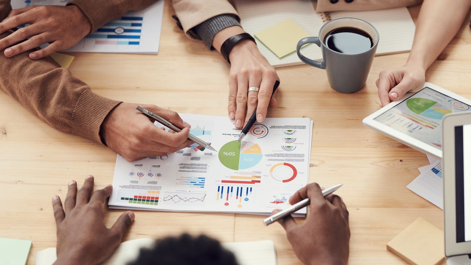 Four people around a desk pointing pens at printed charts beside a tablet and coffee, the client review work accounting compliance automation clears time for