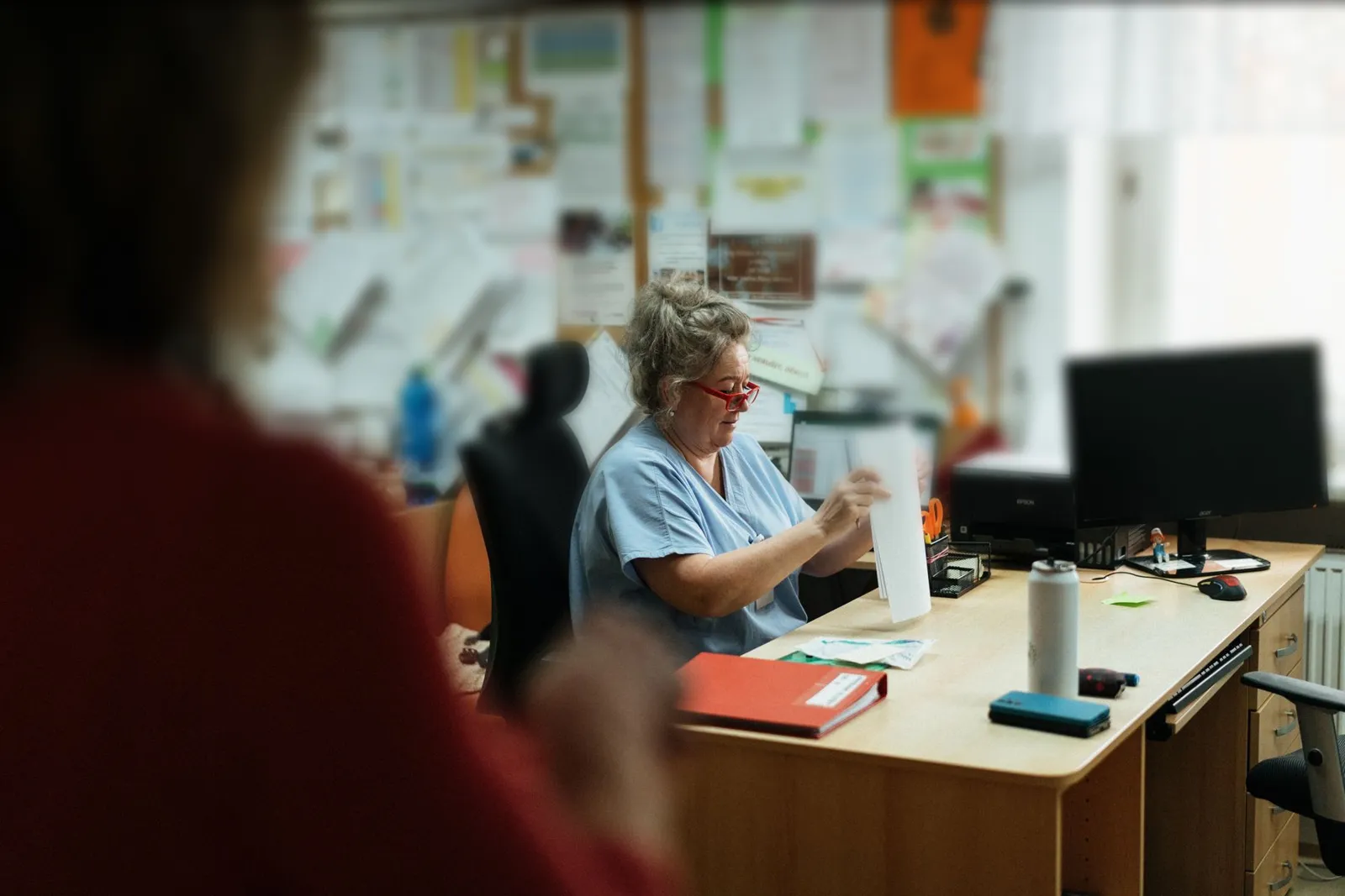 Aged care worker in blue scrubs at an office desk under a noticeboard covered in paperwork, the admin that aged care compliance automation is built to take on