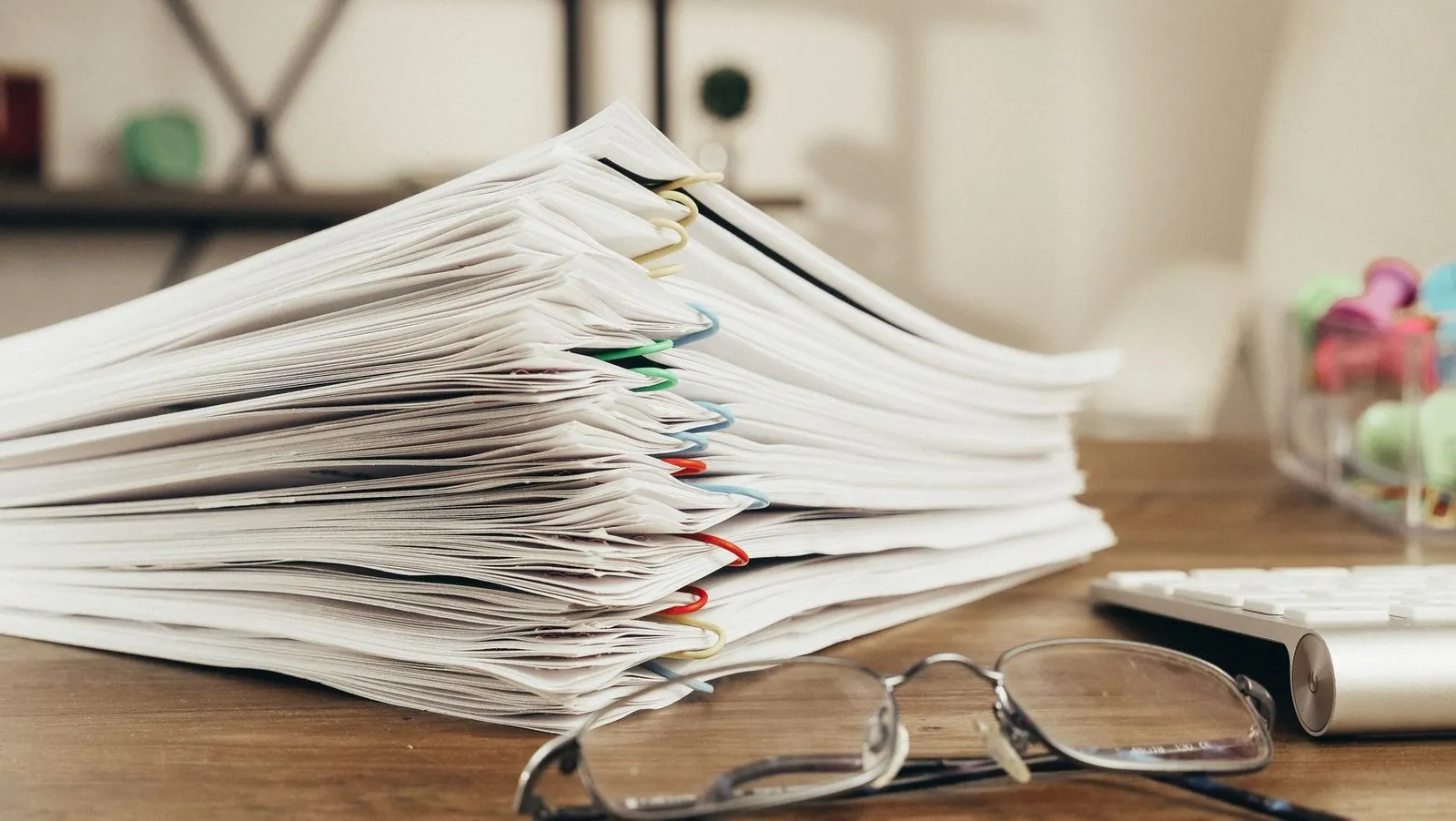 A tall stack of paper clipped documents on a wooden desk with a pair of reading glasses in front