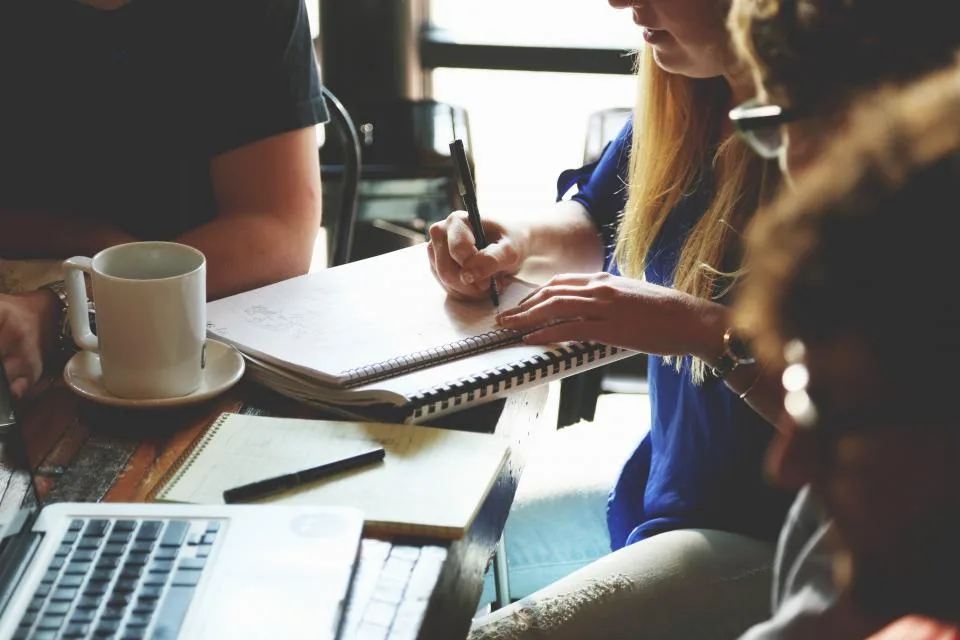 Three colleagues sitting around a wooden table with notebooks, pens and a laptop, reviewing how work moves through the team