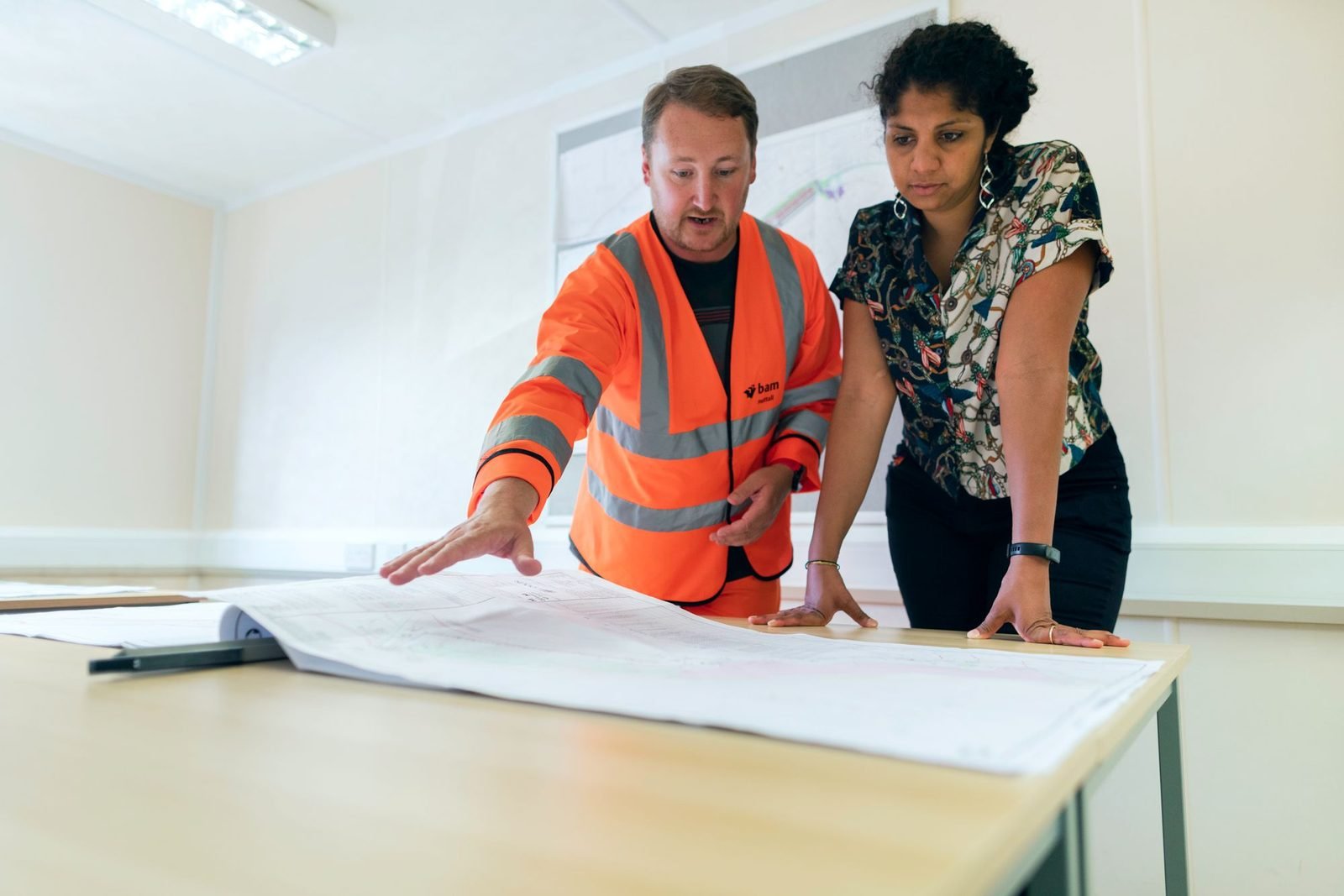 Two engineers in a site office leaning over a large blueprint spread across a table