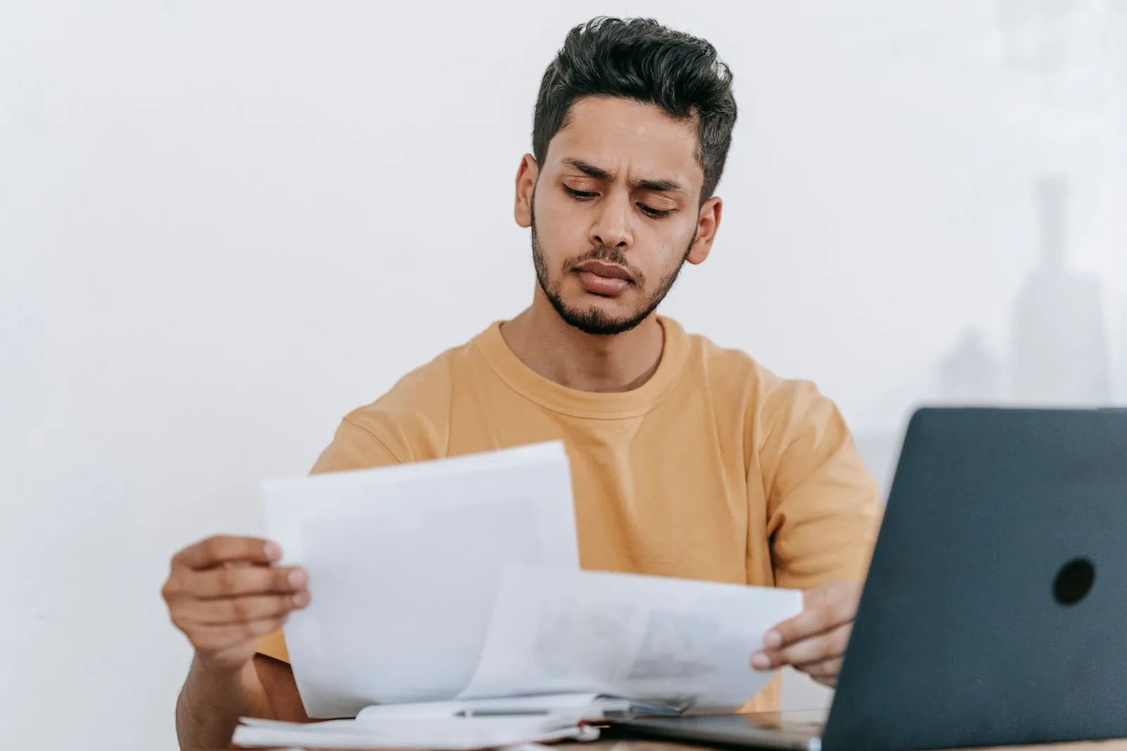 A young man in a mustard T-shirt frowning at printed documents beside an open laptop