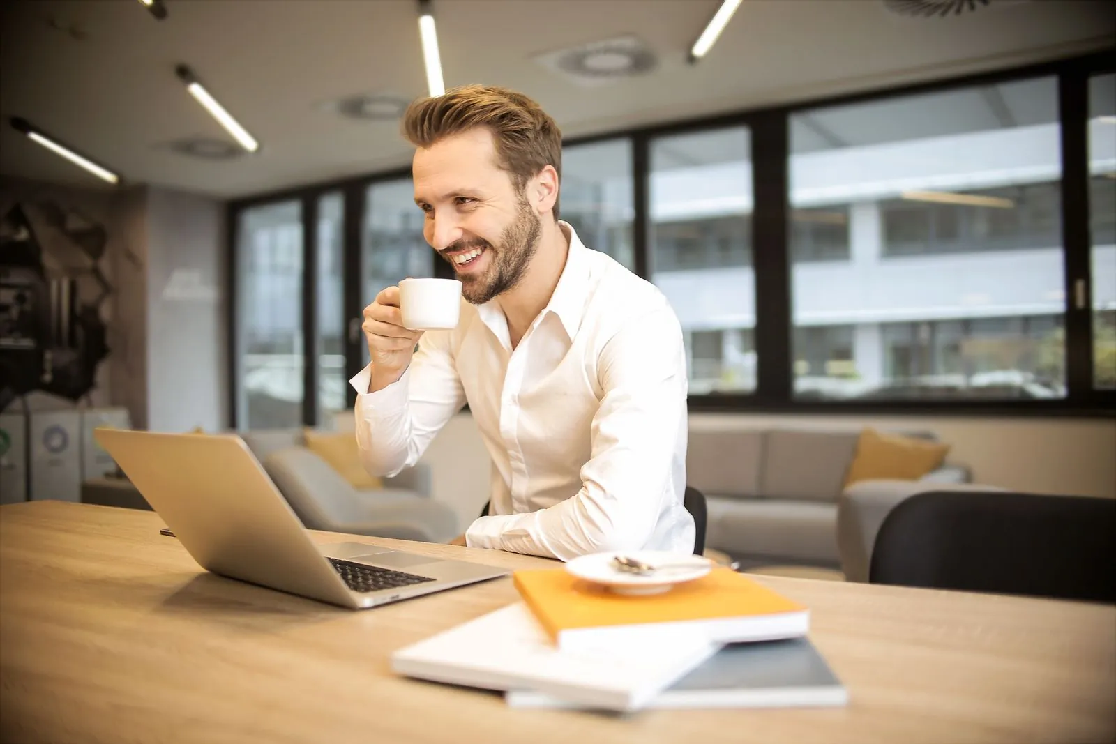 A smiling man sips coffee at a tidy wooden desk with an open laptop and notebooks in a bright office