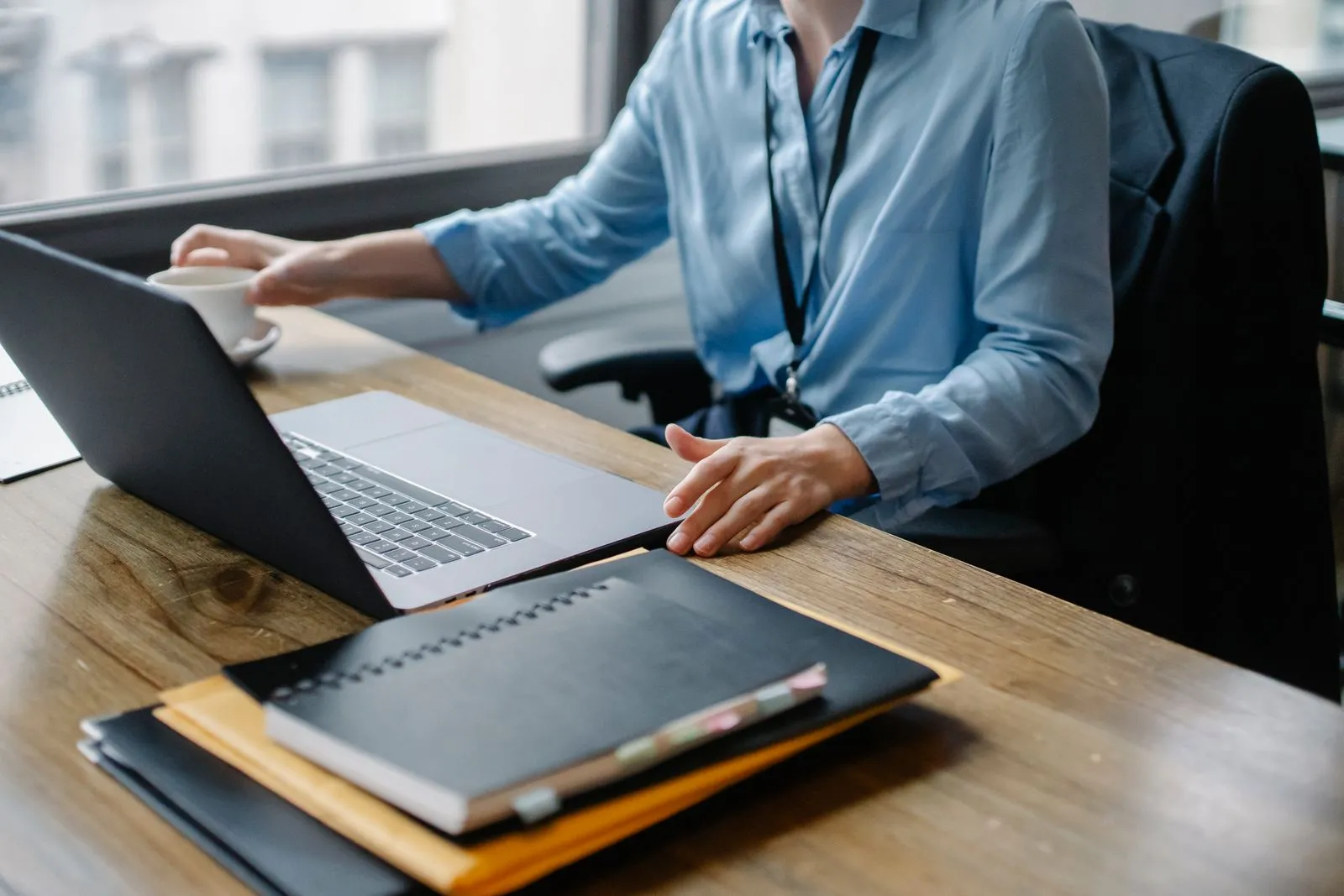 A person in a light blue shirt holding a coffee cup beside an open laptop on a wooden desk
