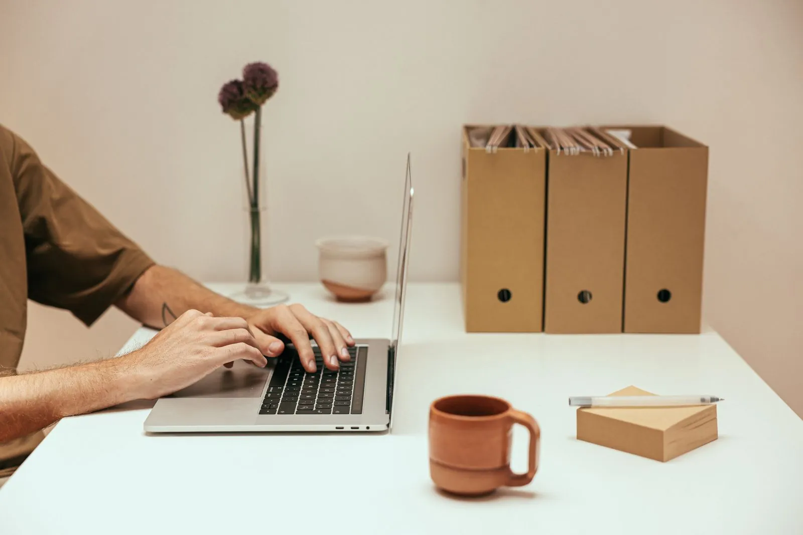 Hands typing on a laptop at a tidy white desk with a terracotta mug, a dried flower and kraft file boxes