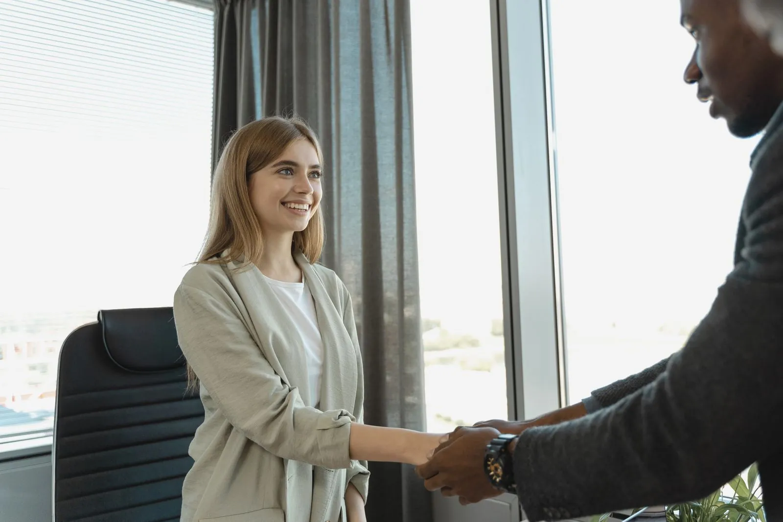 A smiling candidate in a beige blazer shaking hands with an interviewer by a bright office window