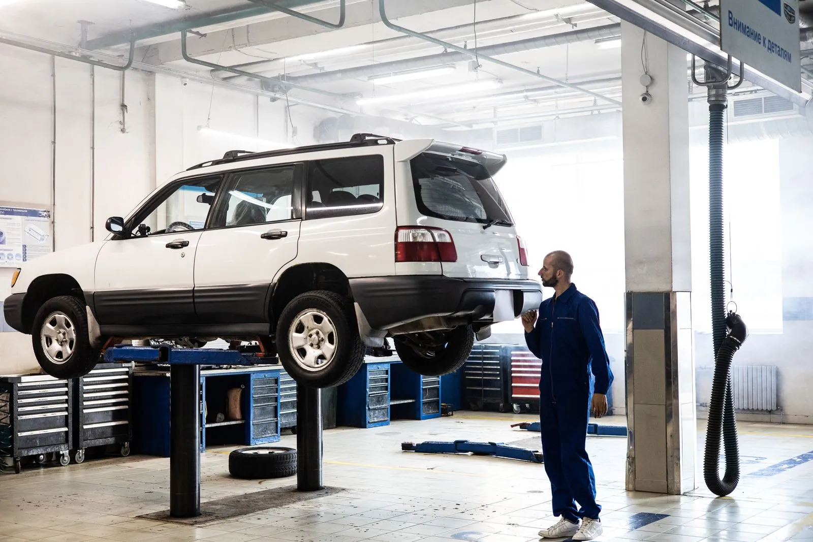 A white SUV raised on a hoist in a bright workshop while a mechanic in blue overalls looks up at it