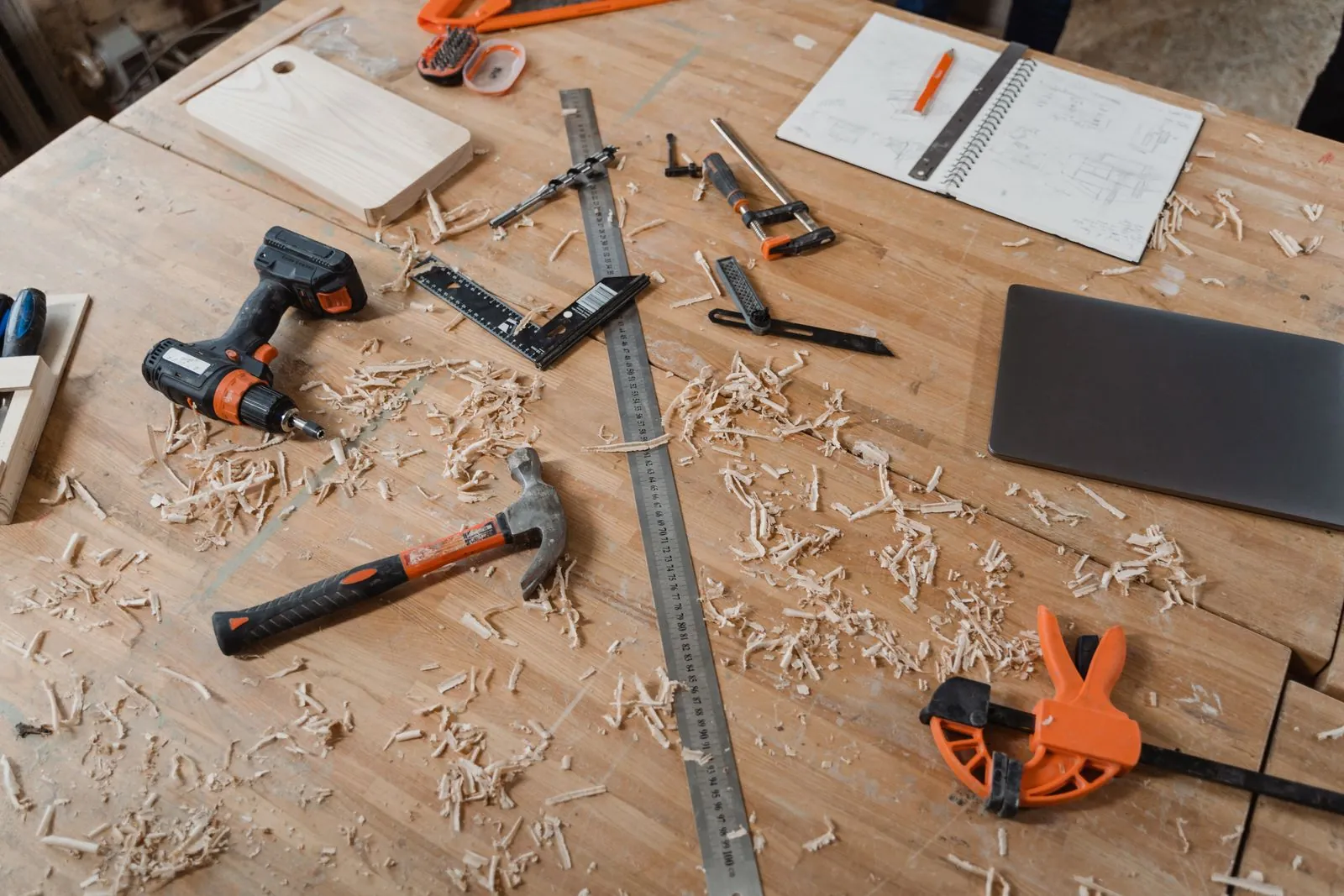 A timber workbench with wood shavings, a hammer, a cordless drill, a steel ruler, an orange clamp and a notebook of sketches