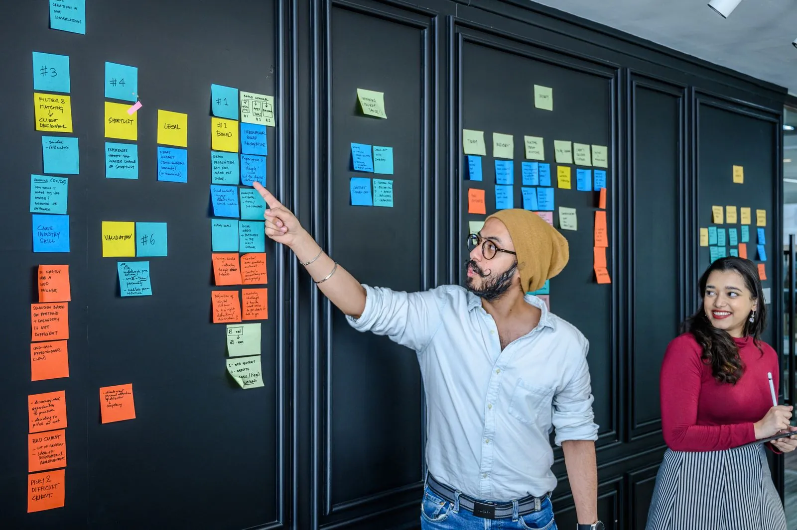 A man points at columns of blue, orange and yellow sticky notes on a dark office wall while a smiling colleague looks on