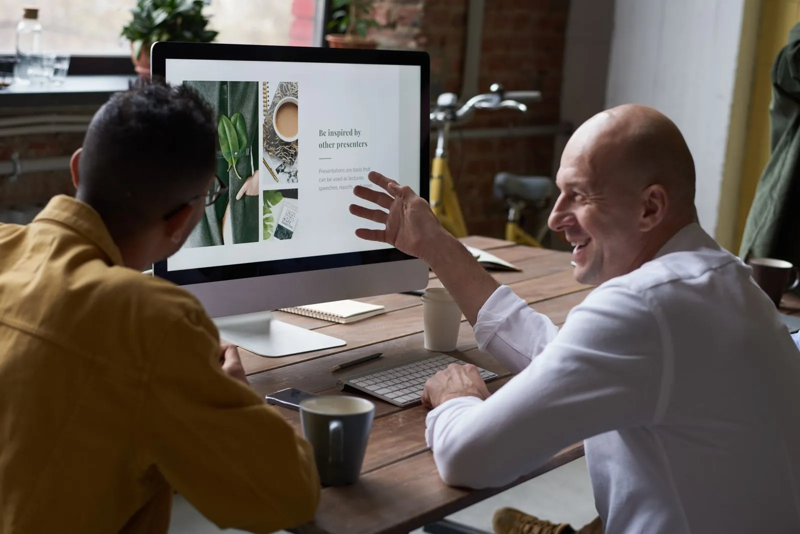 Two colleagues at a wooden desk talking through something on a computer screen, one gesturing at it