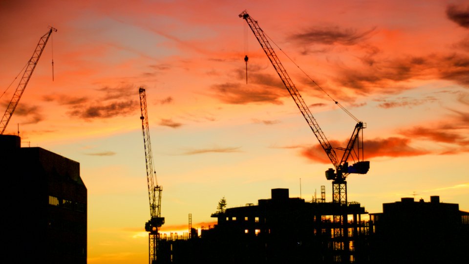 Tower cranes silhouetted against an orange sunset over a half built apartment block