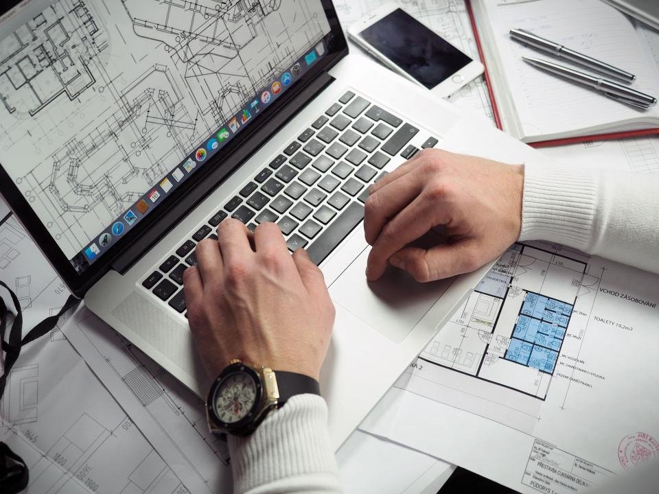 Hands working on a laptop showing architectural drawings, with printed floor plans spread across the desk