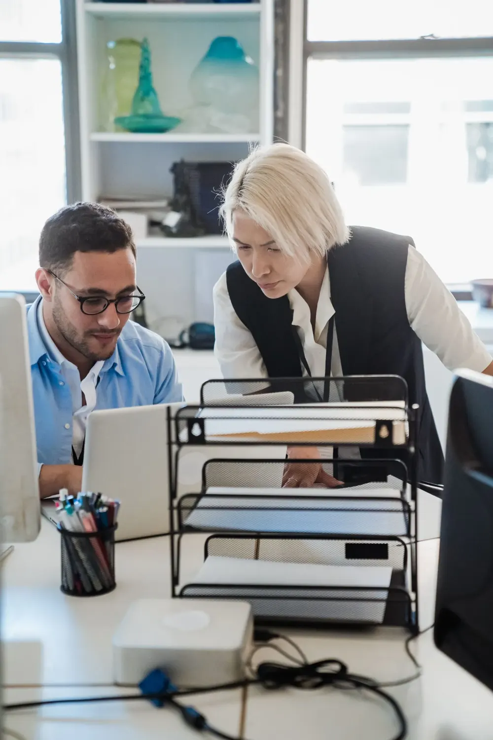 An AI consultant leans over a desk to look at a colleague's laptop screen in a bright open plan office