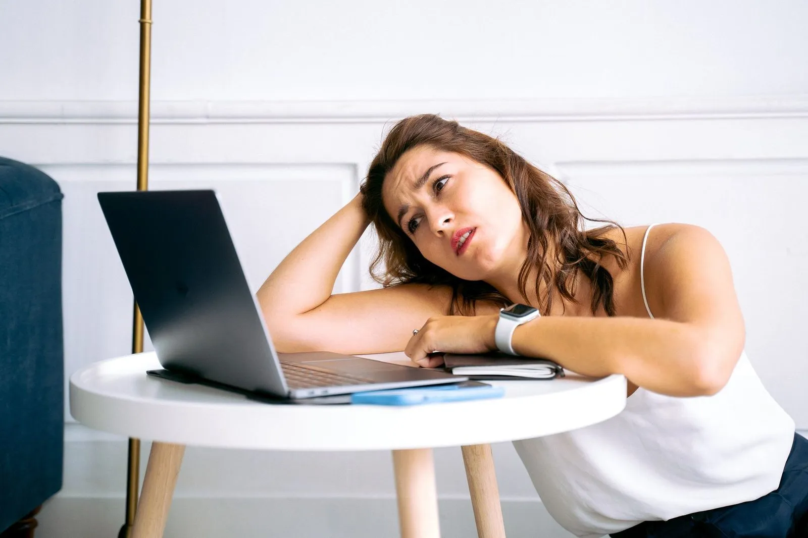 A tired woman rests her head on her hand and stares at an open laptop on a small white table