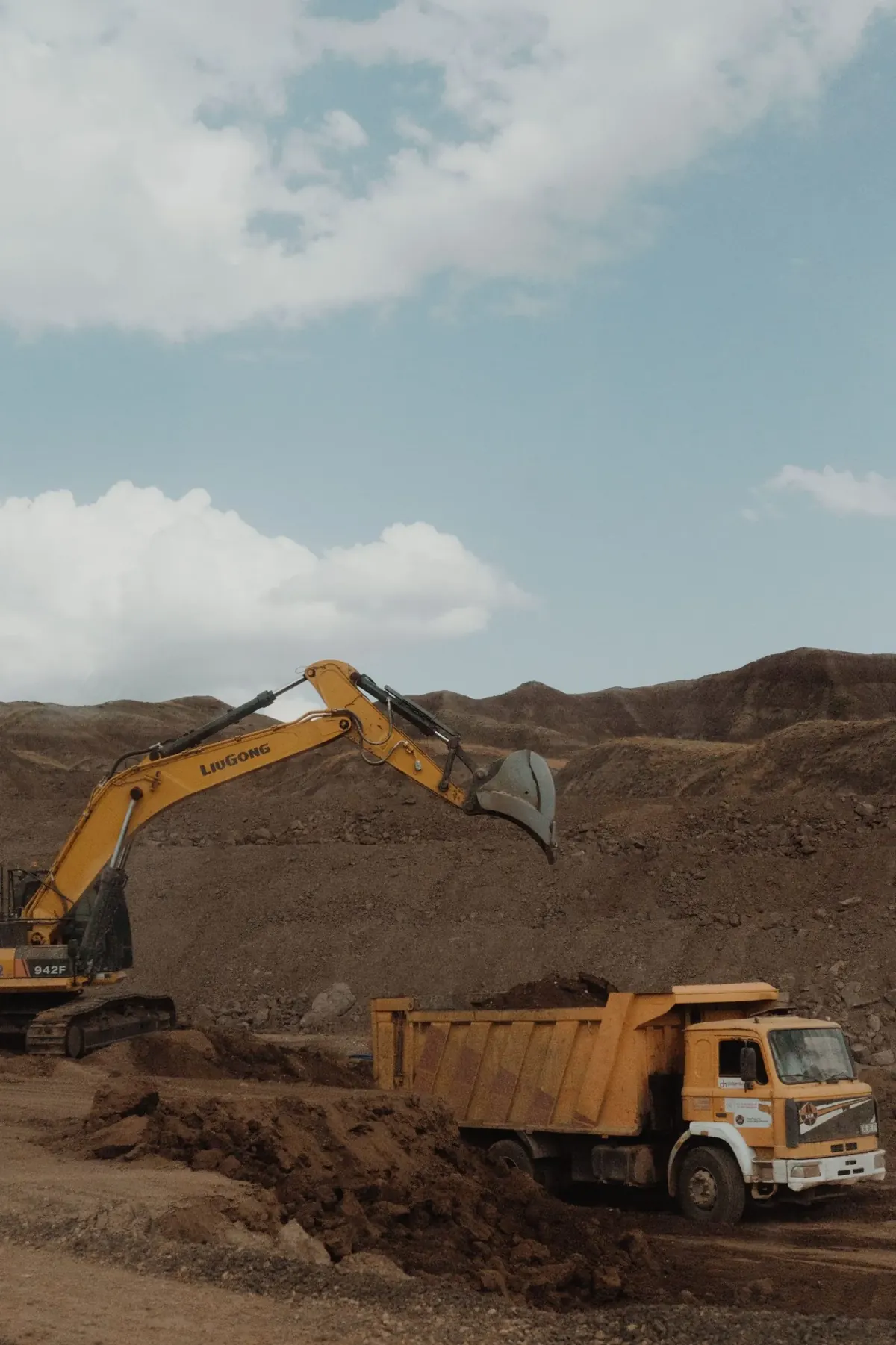 A yellow excavator loading earth into a tipper truck at a quarry under a blue sky