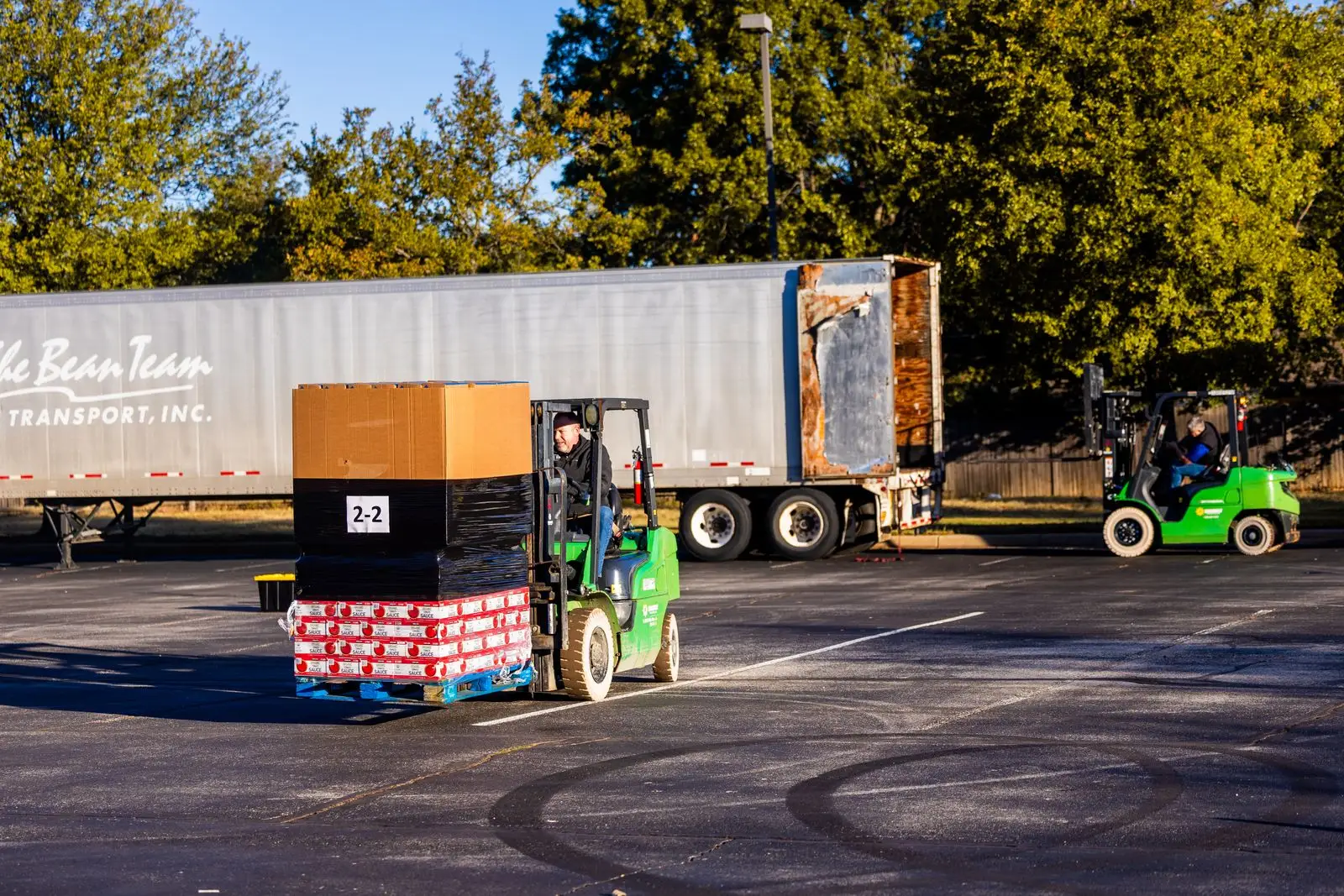 Two forklifts carrying wrapped pallets towards a semi-trailer in a sunny yard