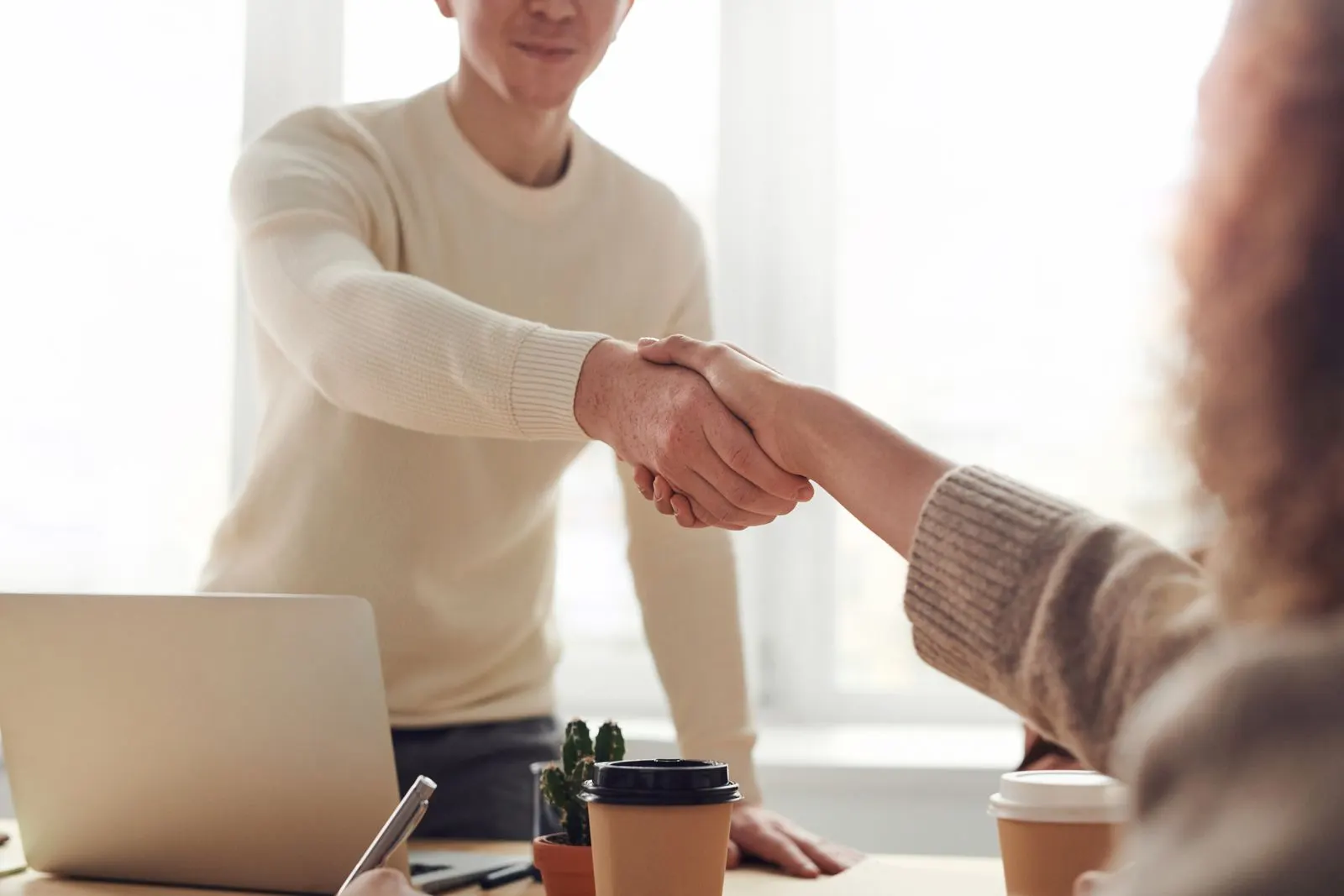 Two professionals shaking hands over a desk with a laptop and takeaway coffee cups after agreeing a fixed-price AI consulting scope