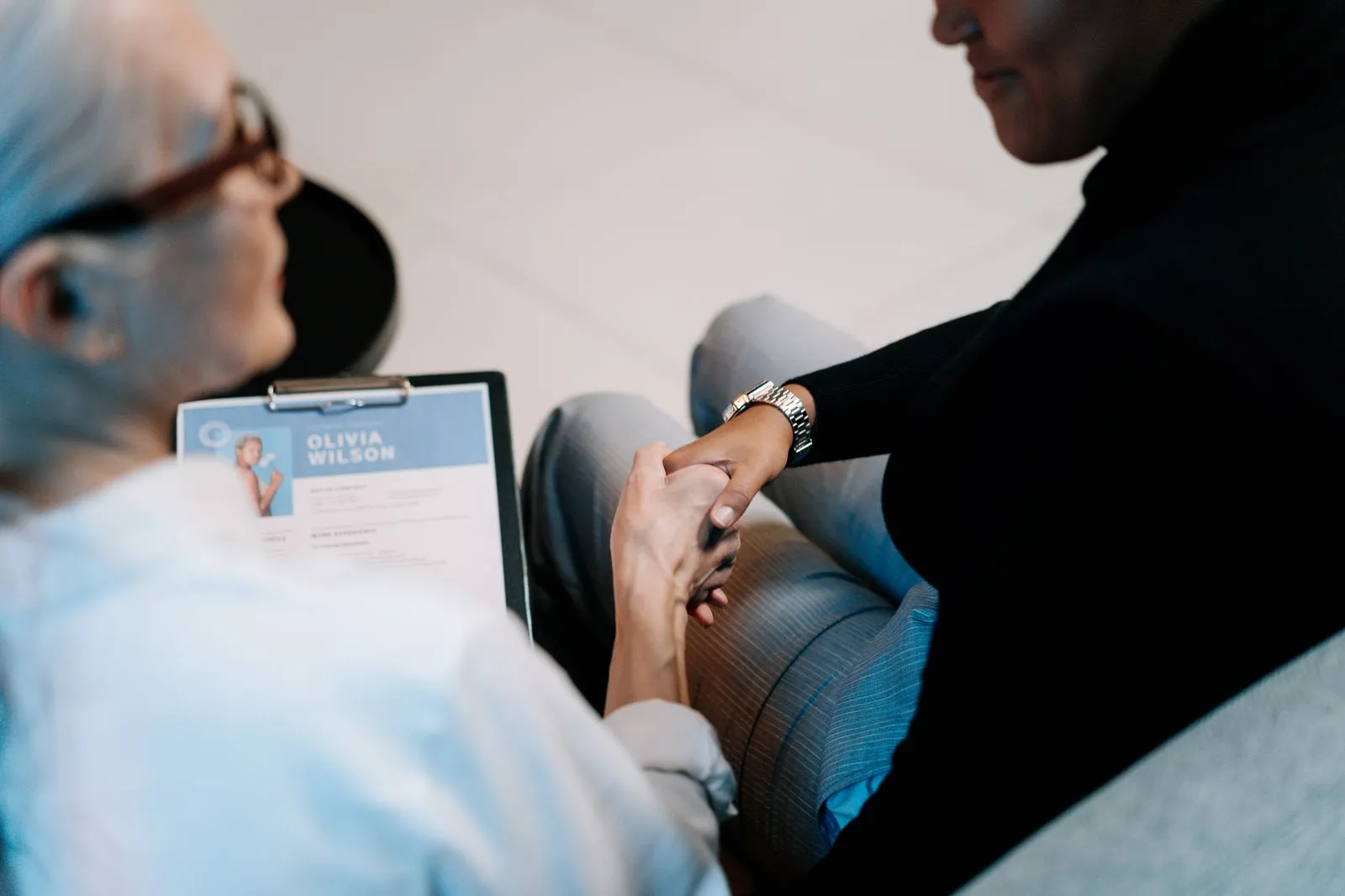 Two women shake hands at a job interview while one holds a clipboard with a printed résumé