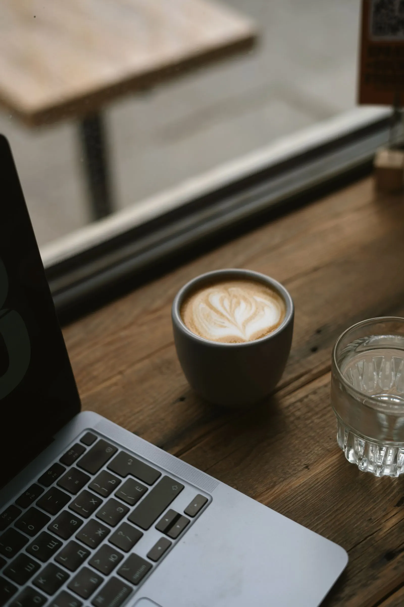A laptop and a latte with leaf art on a wooden cafe table by a window