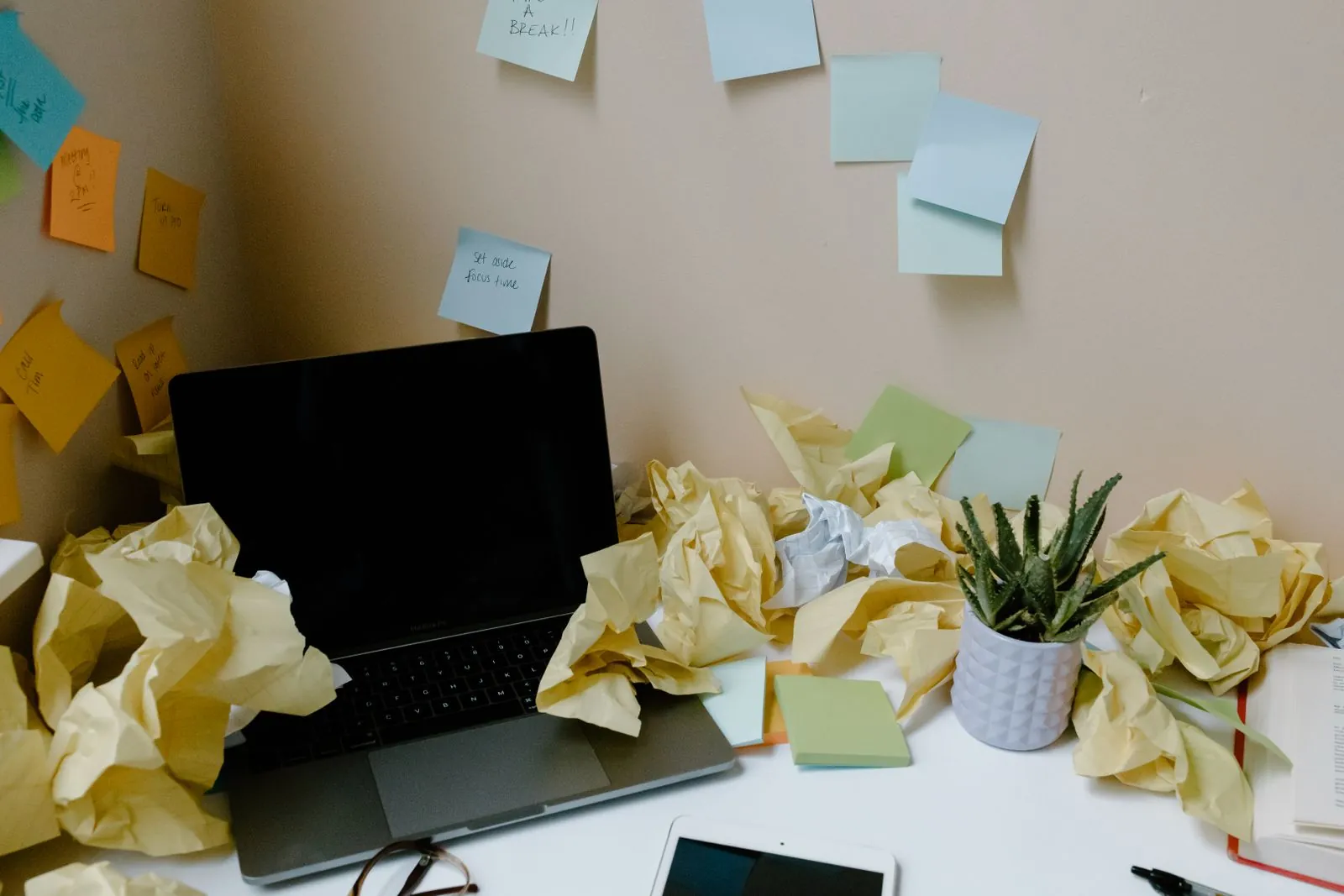 A laptop on a desk surrounded by crumpled yellow paper and sticky notes on the wall behind