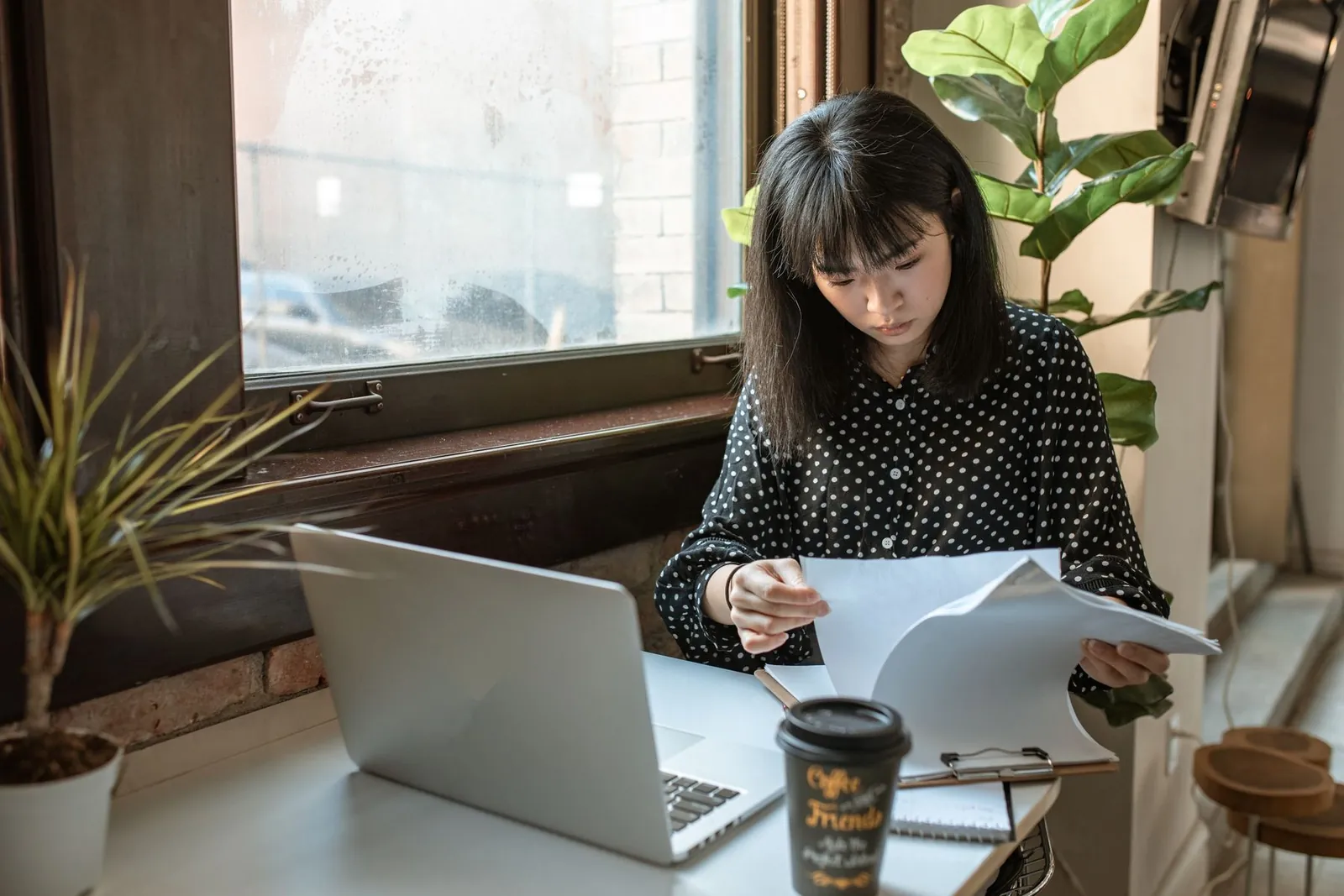 A woman at a laptop leafs through printed pages beside a takeaway coffee cup, checking documents against the screen