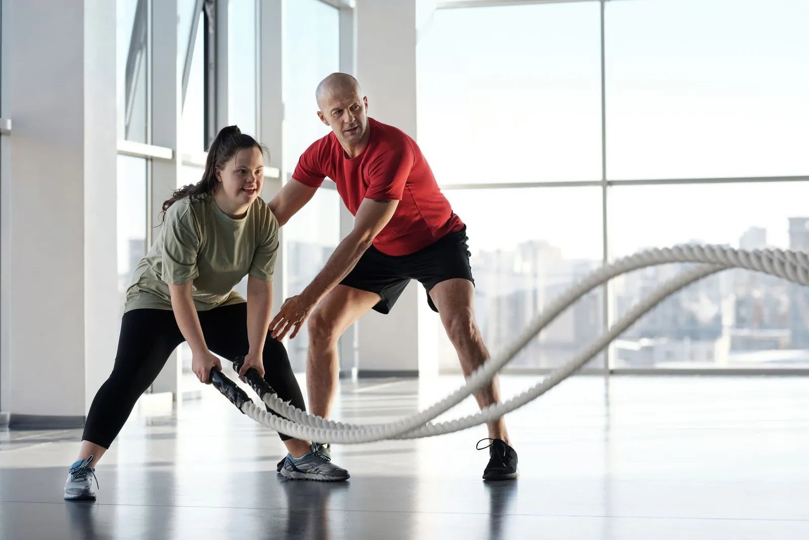 A personal trainer in a red shirt coaches a woman through a battle rope exercise in a bright gym with city views