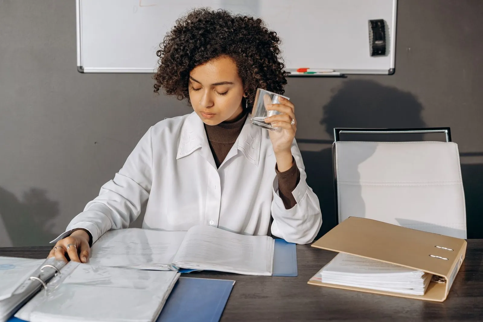 A woman at an office desk reading through papers in a ring binder while holding a glass of water
