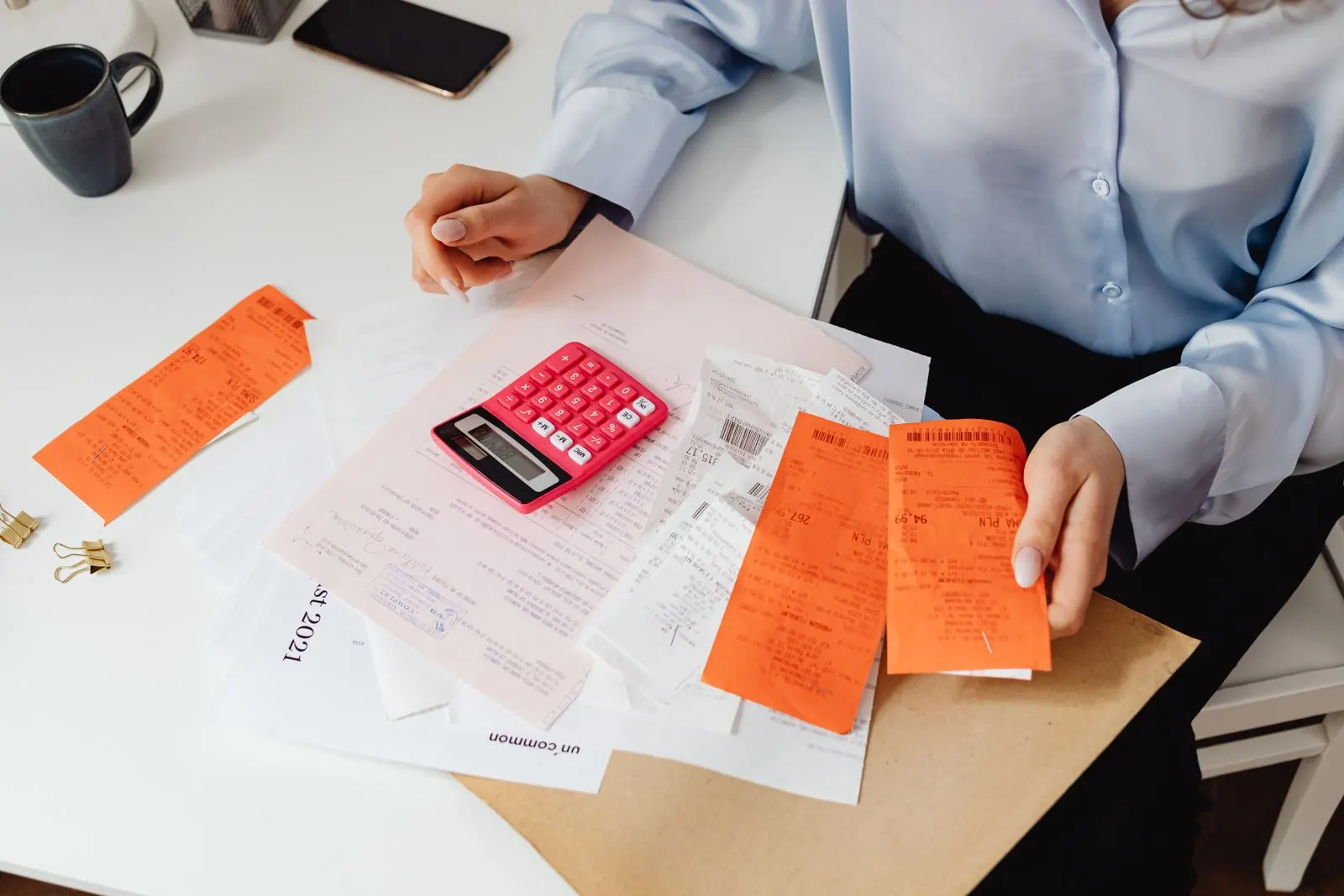 Hands sorting orange receipts beside a pink calculator on a desk, tallying the running costs of an AI automation