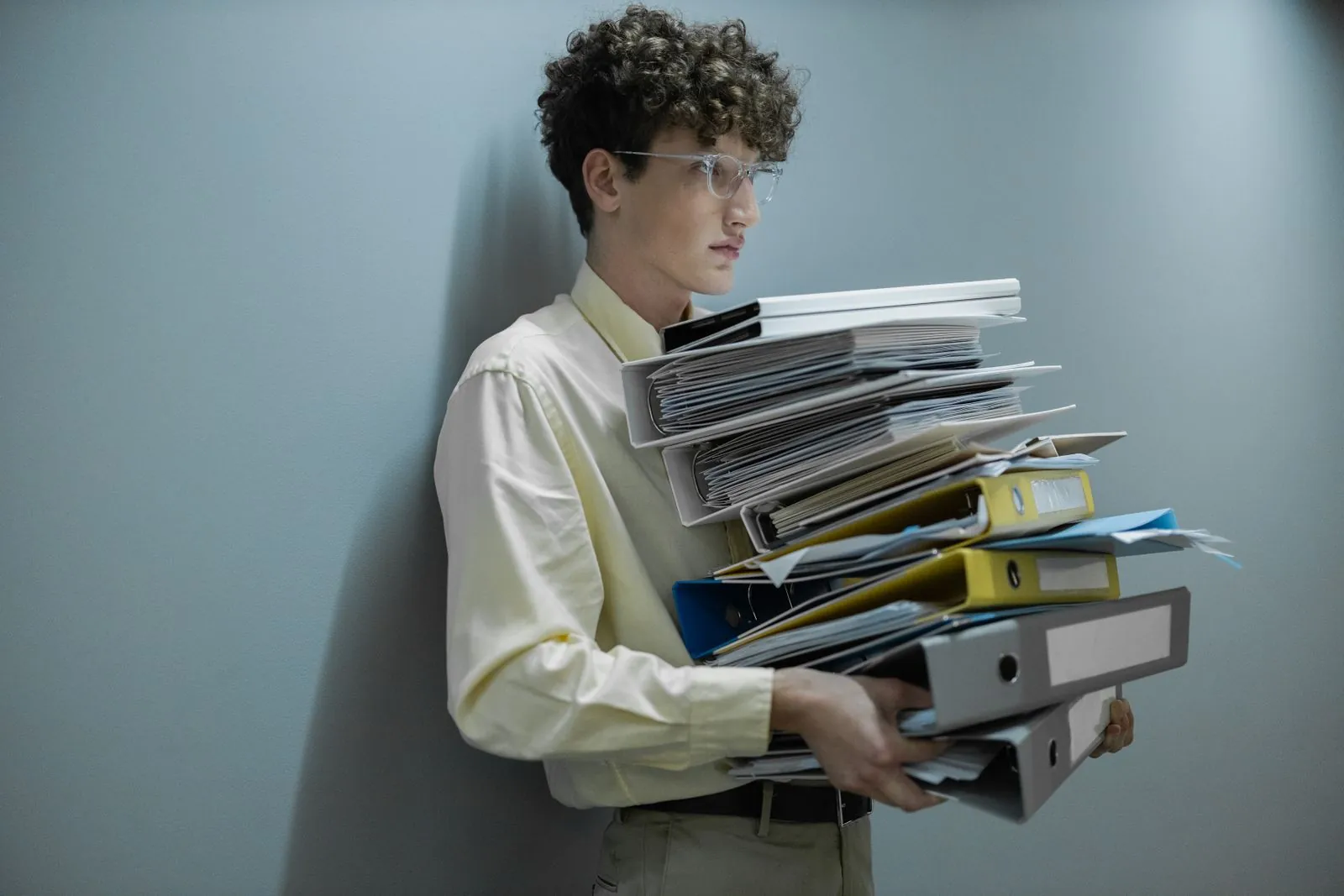 A young man in glasses leaning on a wall, carrying a tall stack of folders and binders