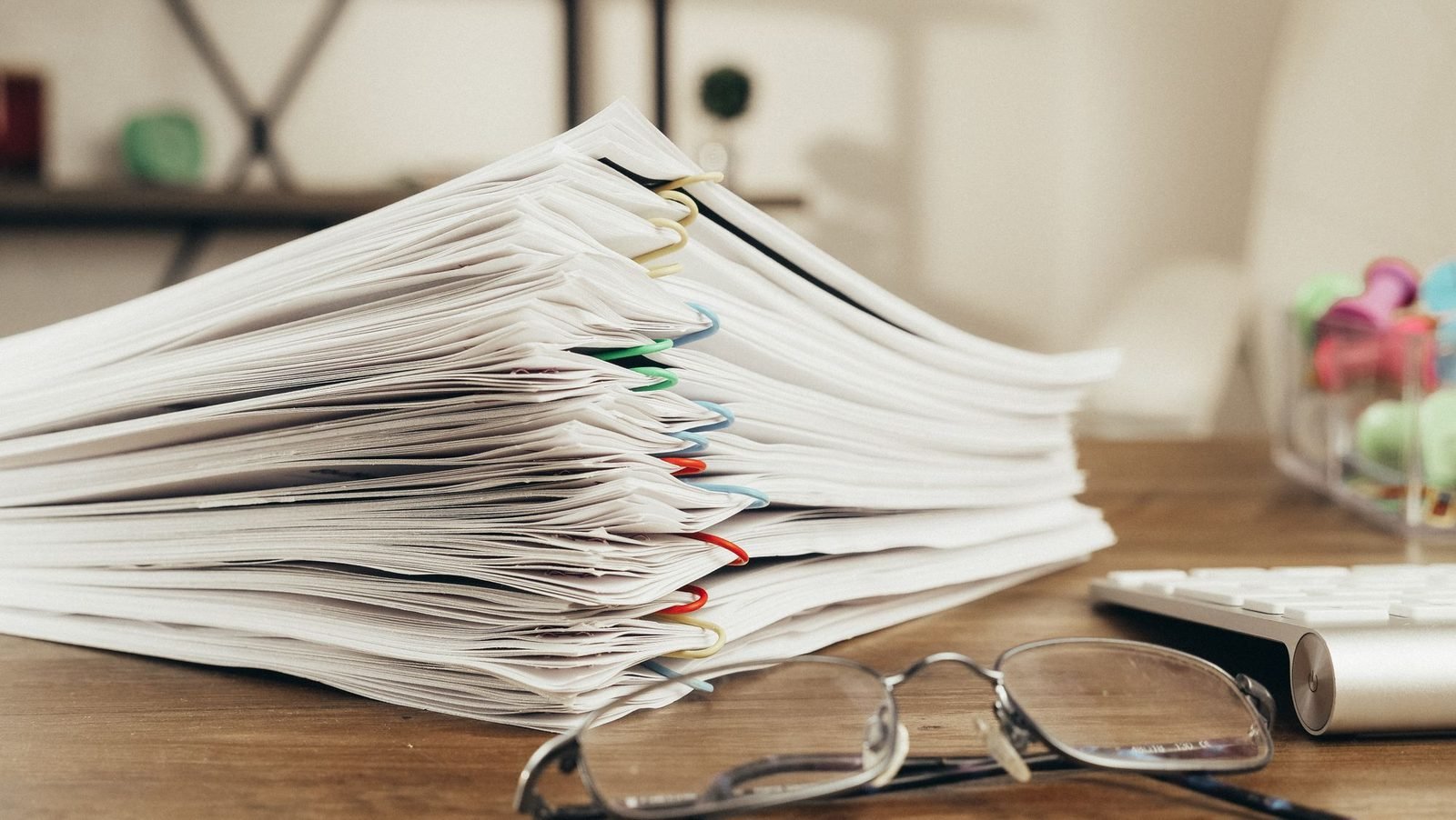 A close-up stack of paper-clipped compliance documents on a desk beside a pair of glasses