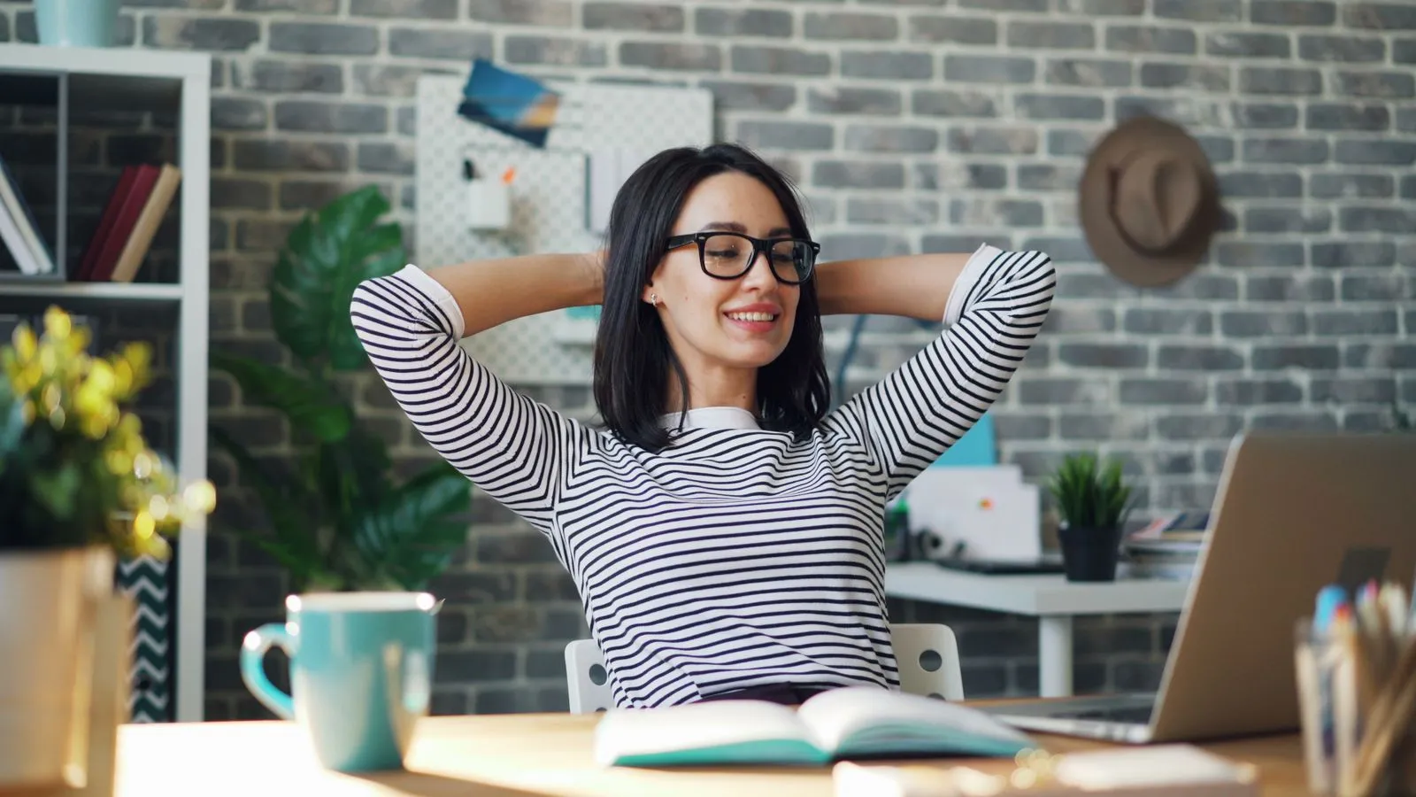 A smiling woman in glasses leans back at her desk with her hands behind her head in front of an open laptop
