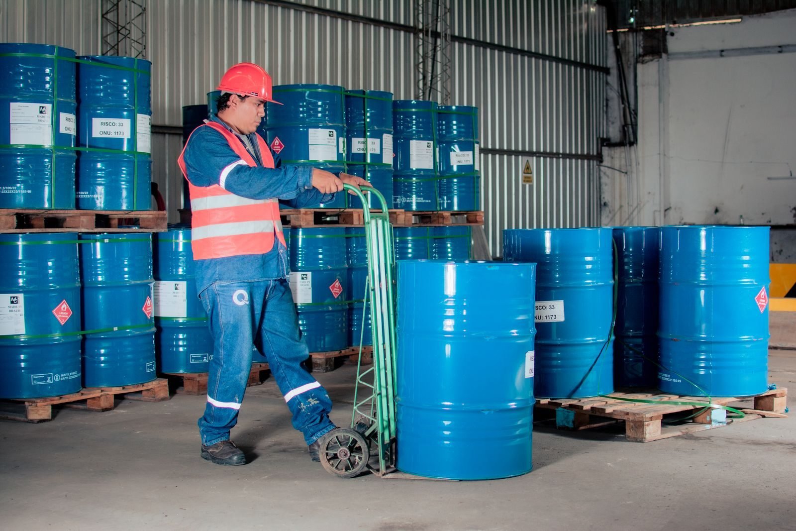 Industrial drums stacked on pallets in a warehouse while a worker in hi-vis moves them, the point where reportable priority waste is classified