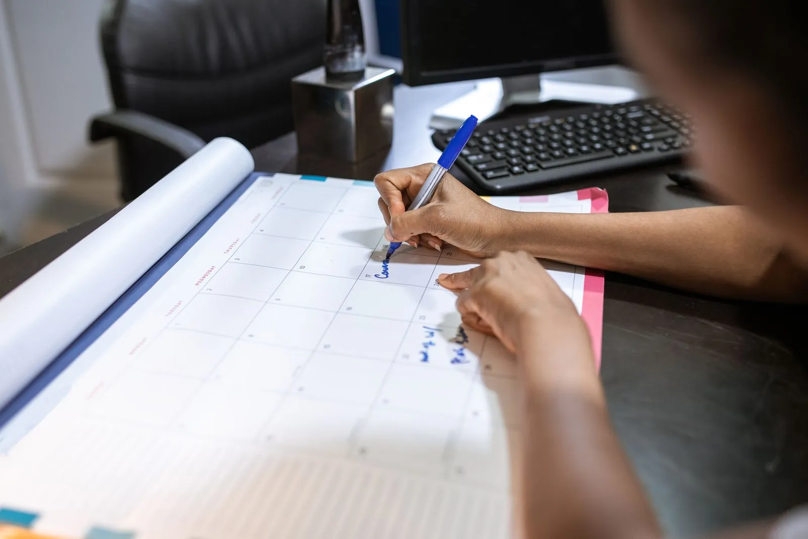 Close-up of a hand writing in blue pen on a large monthly desk-pad calendar next to a keyboard