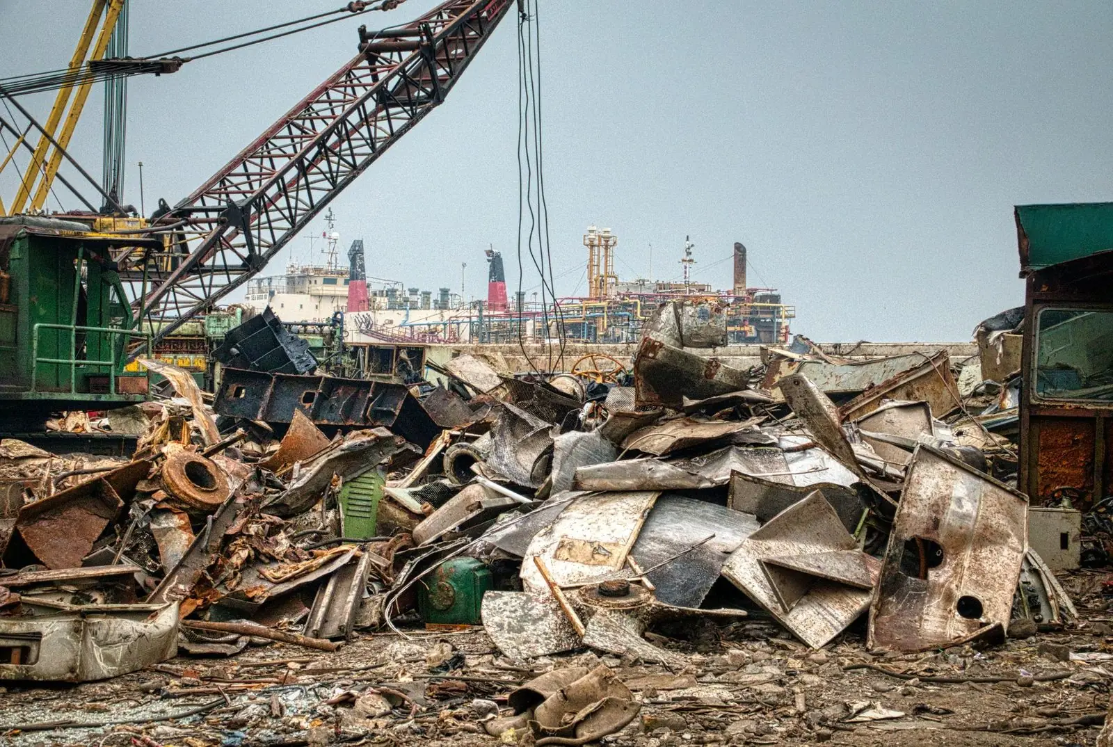 Large piles of rusted scrap metal in a salvage yard with a crane and ships in the background