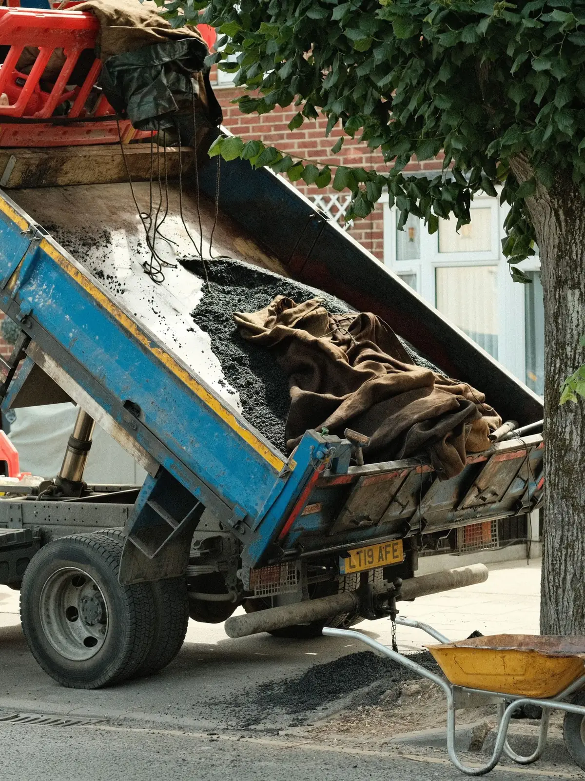 A blue tipper truck unloading broken timber and building rubble beside a tree on a residential street