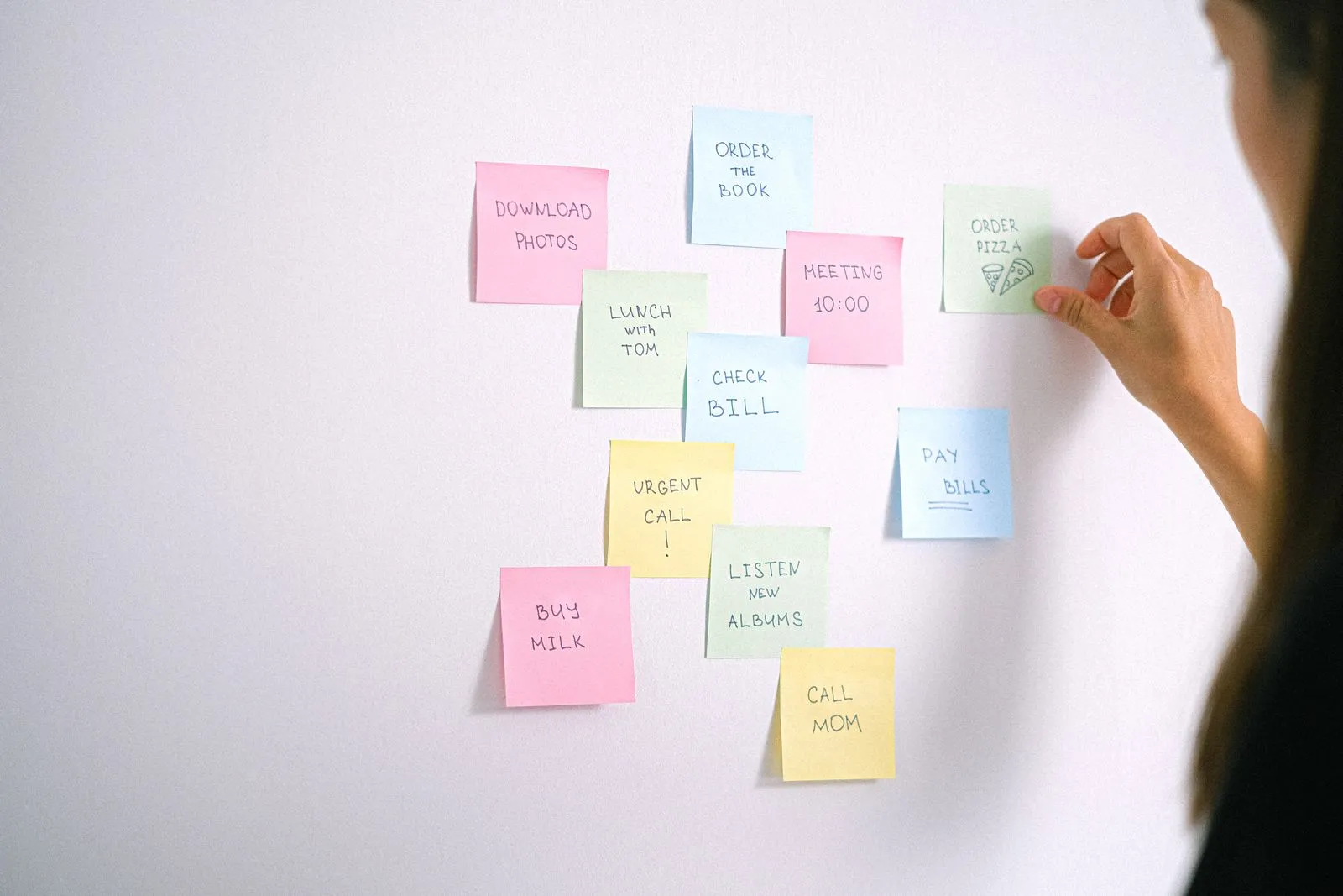 A hand placing pastel sticky notes on a white wall, one reading Urgent call next to one reading Buy milk