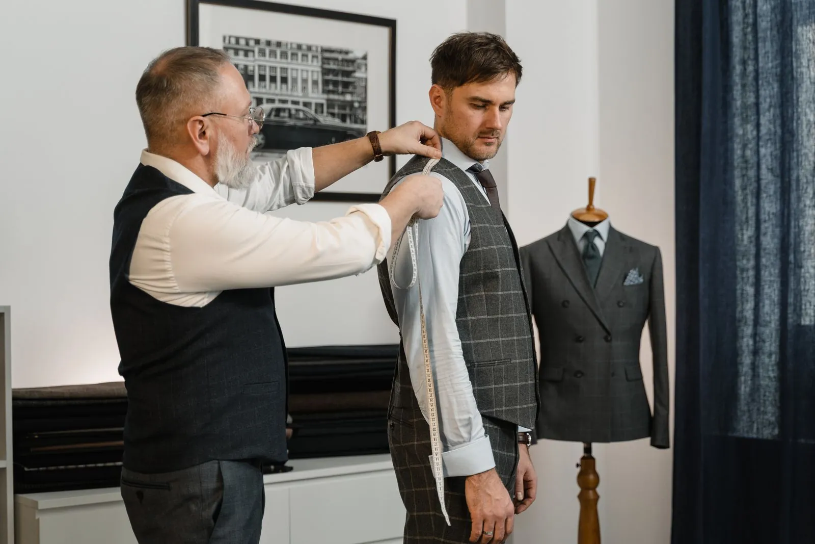 A grey-bearded tailor measures a client's arm with a tape measure beside a suit on a mannequin
