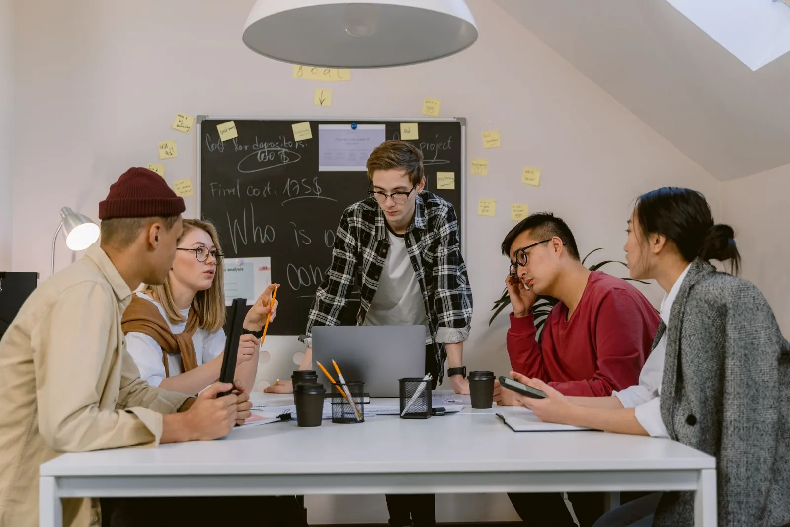 Five colleagues around a white table with a laptop and coffee cups in front of a blackboard covered in sticky notes