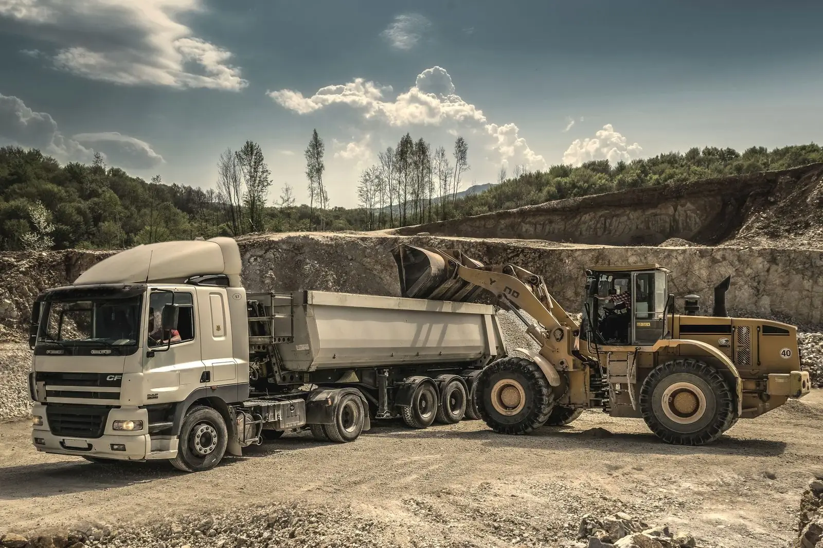 A white tipper truck and a front end loader on a dirt yard under a cloudy sky