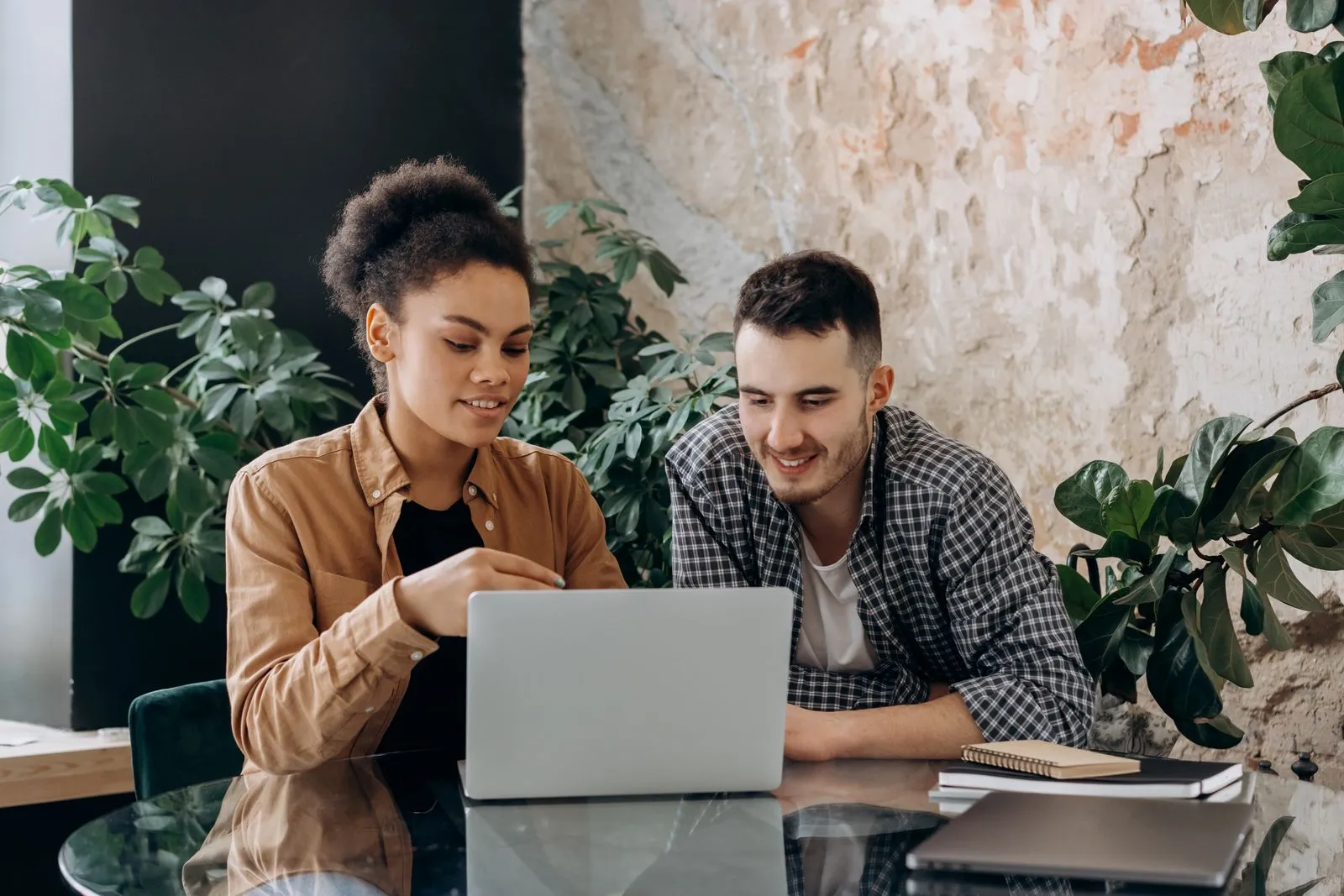 A smiling woman and a man in a check shirt review something together on a silver laptop at a glass table