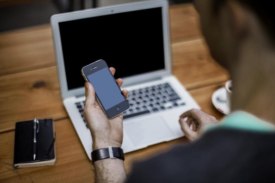 A person holding a mobile phone in front of a laptop, about to call a supplier to verify changed bank details
