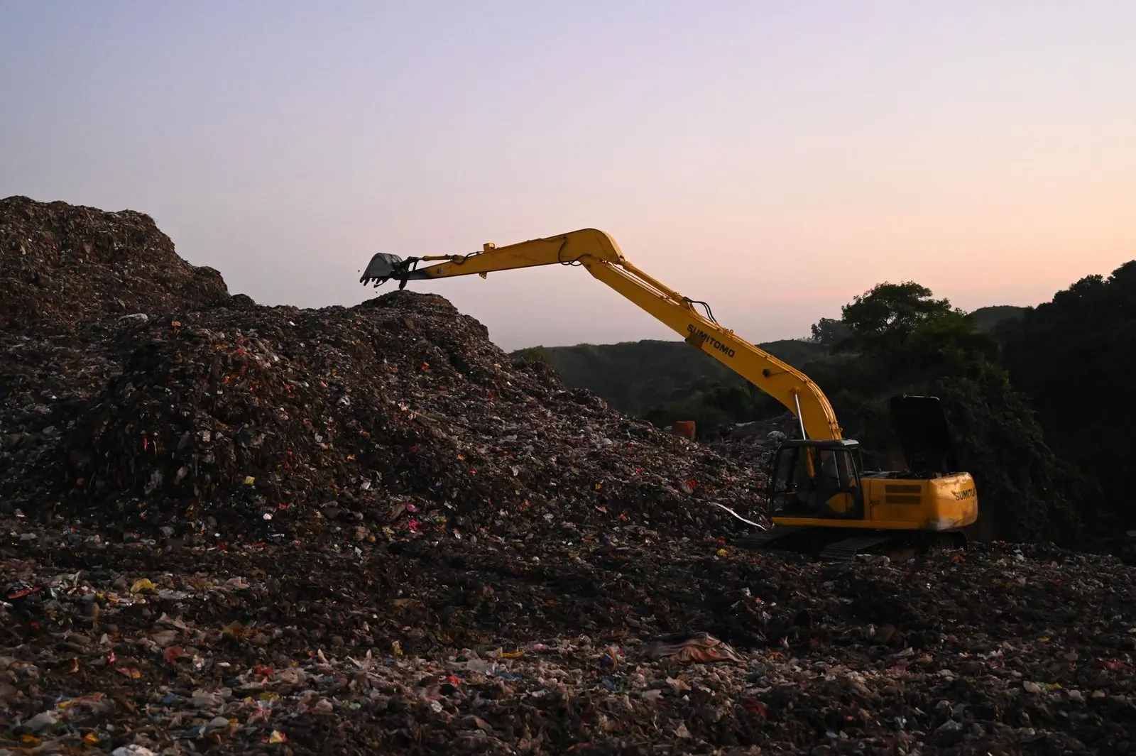 A yellow excavator working a landfill at dusk, the end of the line that waste records automation keeps track of