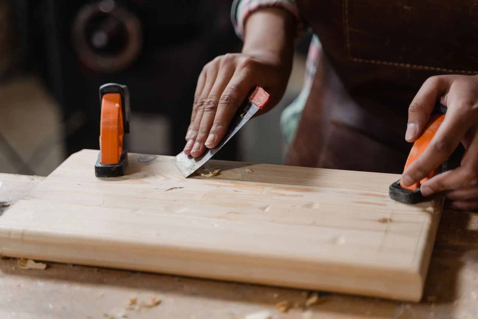 A woodworker's hands scrape glue from a pine board held by orange clamps on a workbench
